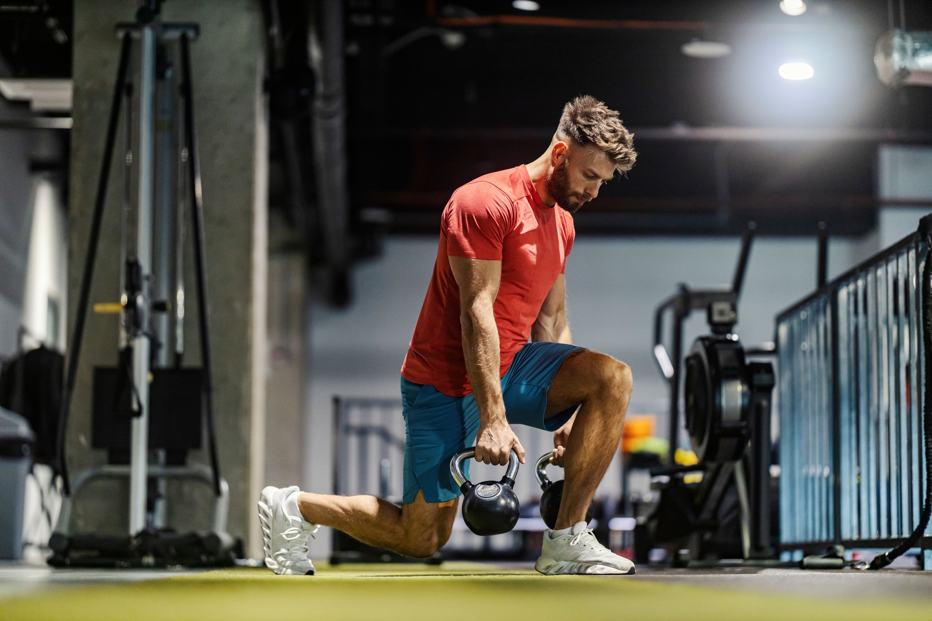 Seitenansicht eines starken, muskulösen Sportlers, der Ausfallschritte macht, während er im Fitnessstudio Kettlebells trägt.