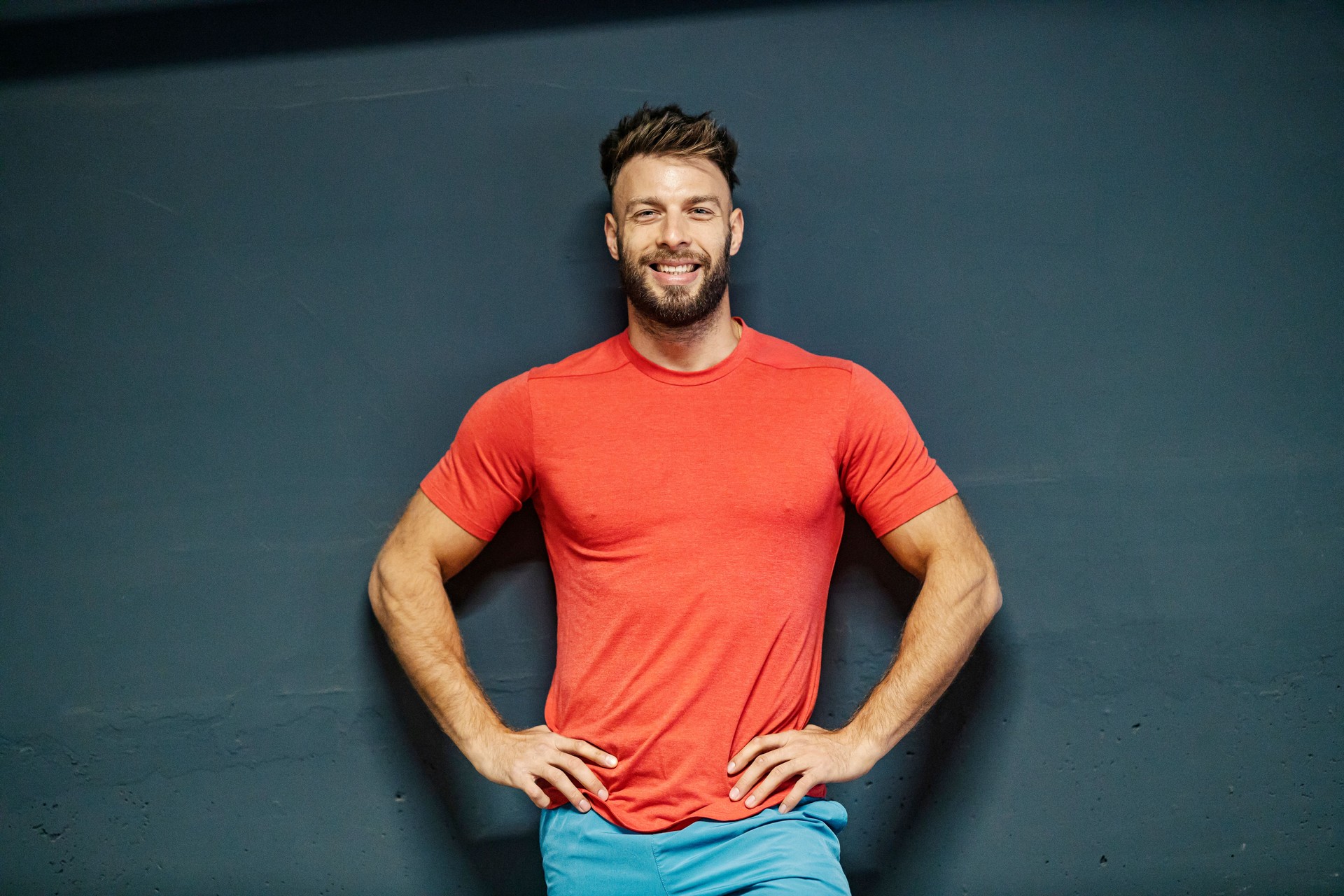 Portrait of a happy healthy fit muscular sportsman standing against the wall with hands on hips and smiling at camera.