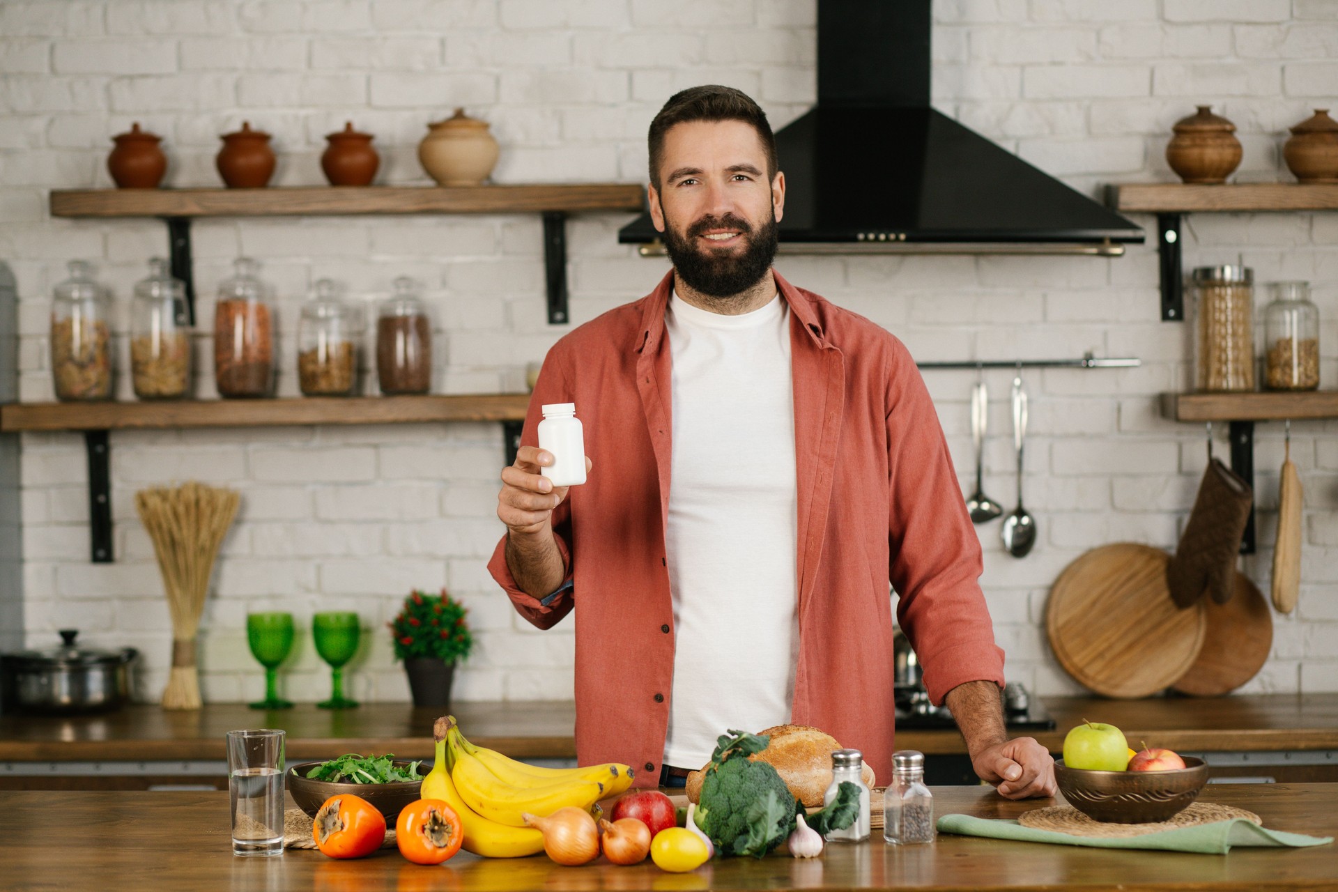 Smiling caucasian man with beard in home kitchen holding bottle of nutritional supplements, looking at the camera.